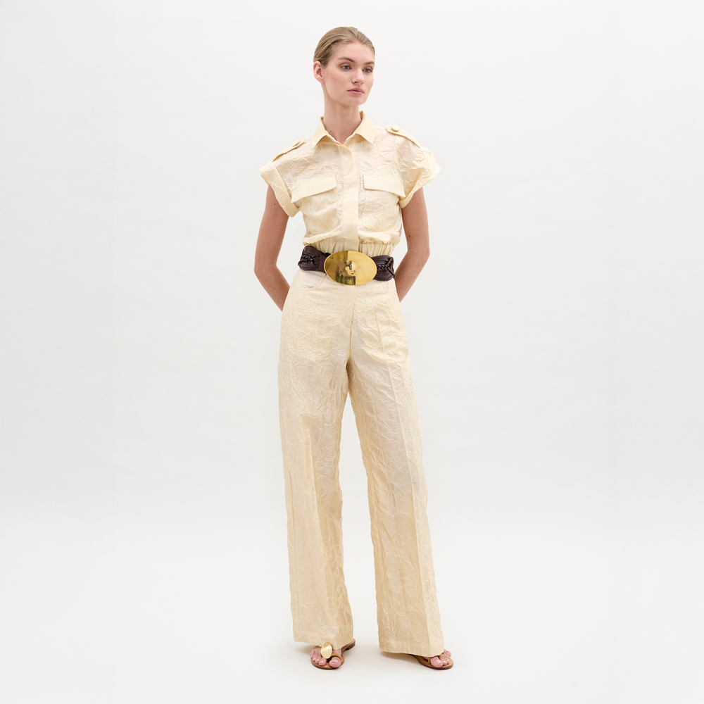 A person stands against a plain background wearing the Emine Pant, a beige textured wide-leg silhouette crafted from Italian fabric, styled with short sleeves, a wide black belt with gold buckle, and sandals.