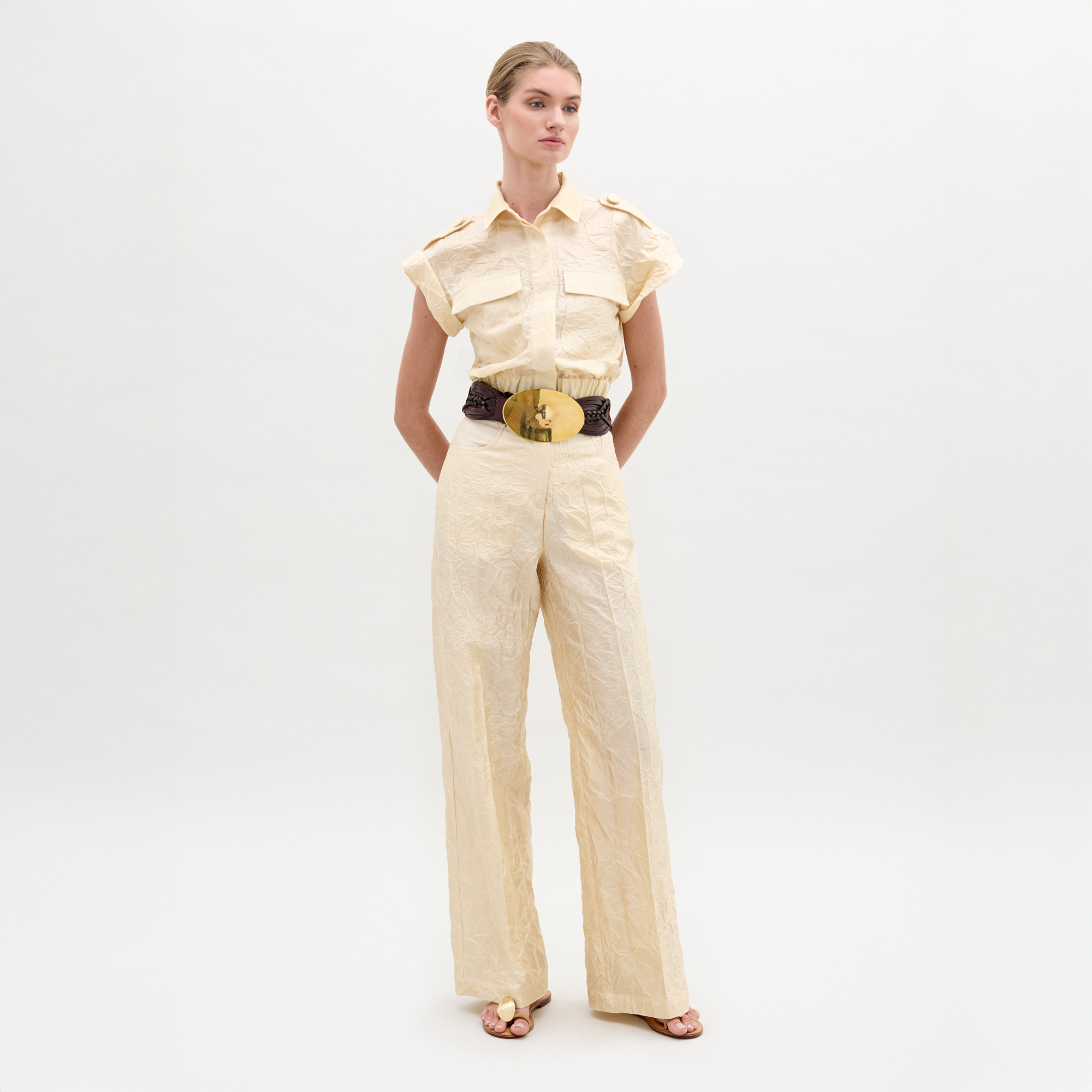 A person stands against a plain background wearing the Emine Pant, a beige textured wide-leg silhouette crafted from Italian fabric, styled with short sleeves, a wide black belt with gold buckle, and sandals.