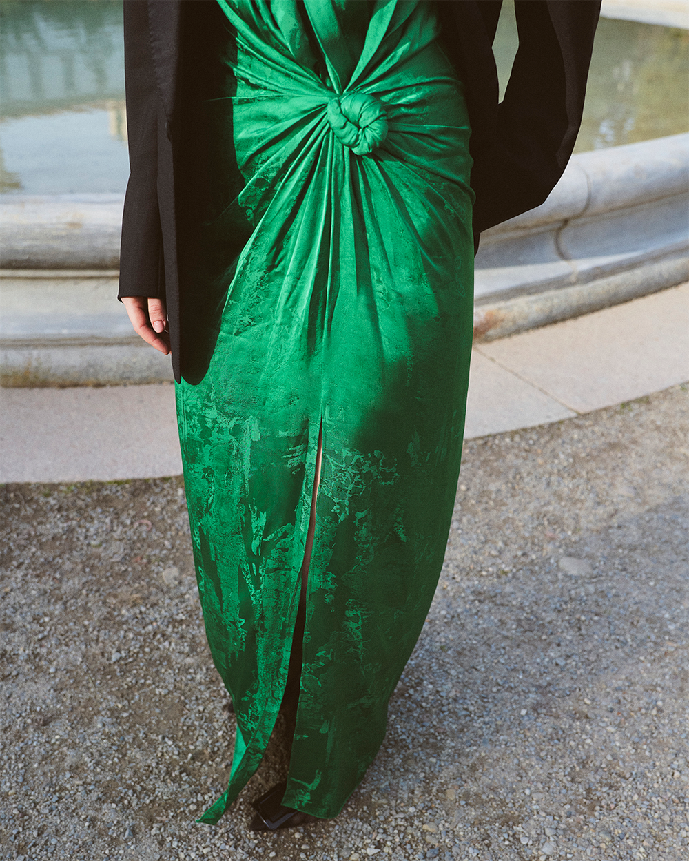 Person wearing a green satin dress with a knot at the waist and a black jacket stands outdoors near a stone water fountain.