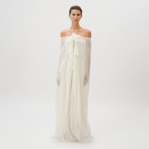 A woman stands against a plain white background, wearing the Edna Dress—an off-the-shoulder, textured white gown crafted from luxurious fabric with long, flowing layers and delicate sheer accents. This exquisite piece is masterfully made in Italy.