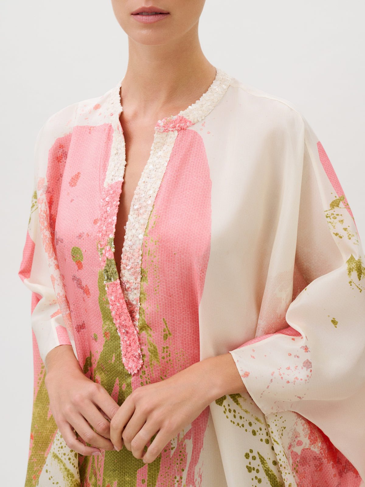 The Elea Tunic is a loose-fitting silk habutai top with a split neckline featuring sequin trim, patterned with abstract pink and green shapes on a cream background.