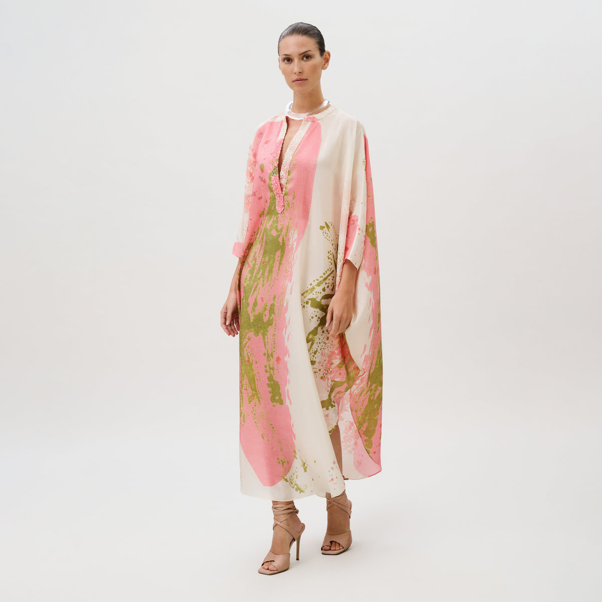 A model wearing an Elea Tunic, a loose, long-sleeved silk top with an abstract pink and green pattern, paired with lace-up heels.