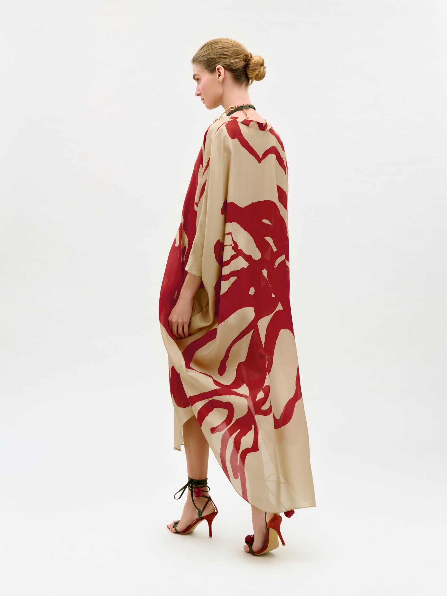 A woman with her hair in a bun wears the Elea Tunic—a loose, beige and red patterned maxi dress made from 100% Italian silk—paired with red high-heeled sandals. She stands against a plain, light background, facing slightly away.