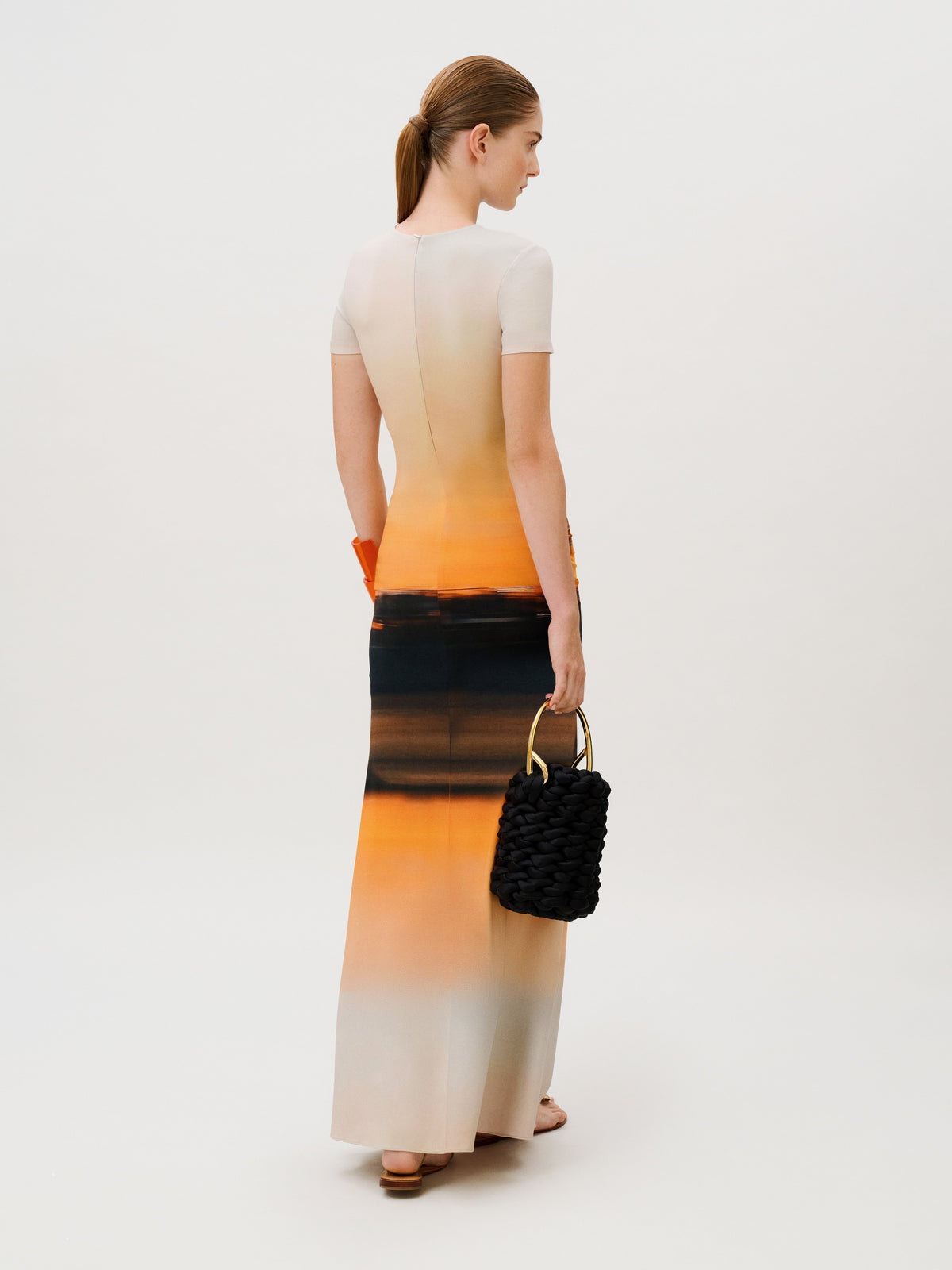 A woman wearing the Elora Dress, a slim-fit, floor-length viscose piece with a cream, orange, and dark charcoal gradient print, carrying a black braided handbag with a gold handle.