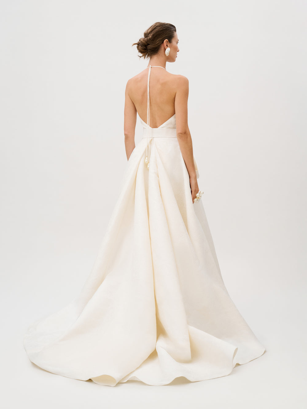 A woman stands facing away, holding a small bouquet, wearing the Elsa Dress—a white halter-neck, backless gown crafted from luxurious Italian fabric with a long train.