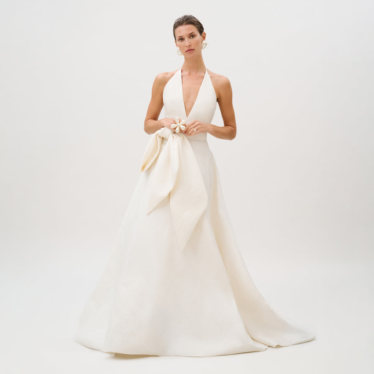Dressed in the exquisite Elsa Dress—a sleeveless, floor-length white gown from our Atelier collection, crafted from luxurious Italian fabric and featuring a deep V neckline with a large bow at the waist—the woman stands against a plain light background.