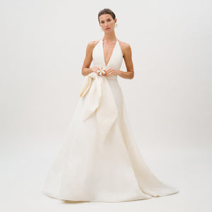 Dressed in the exquisite Elsa Dress—a sleeveless, floor-length white gown from our Atelier collection, crafted from luxurious Italian fabric and featuring a deep V neckline with a large bow at the waist—the woman stands against a plain light background.