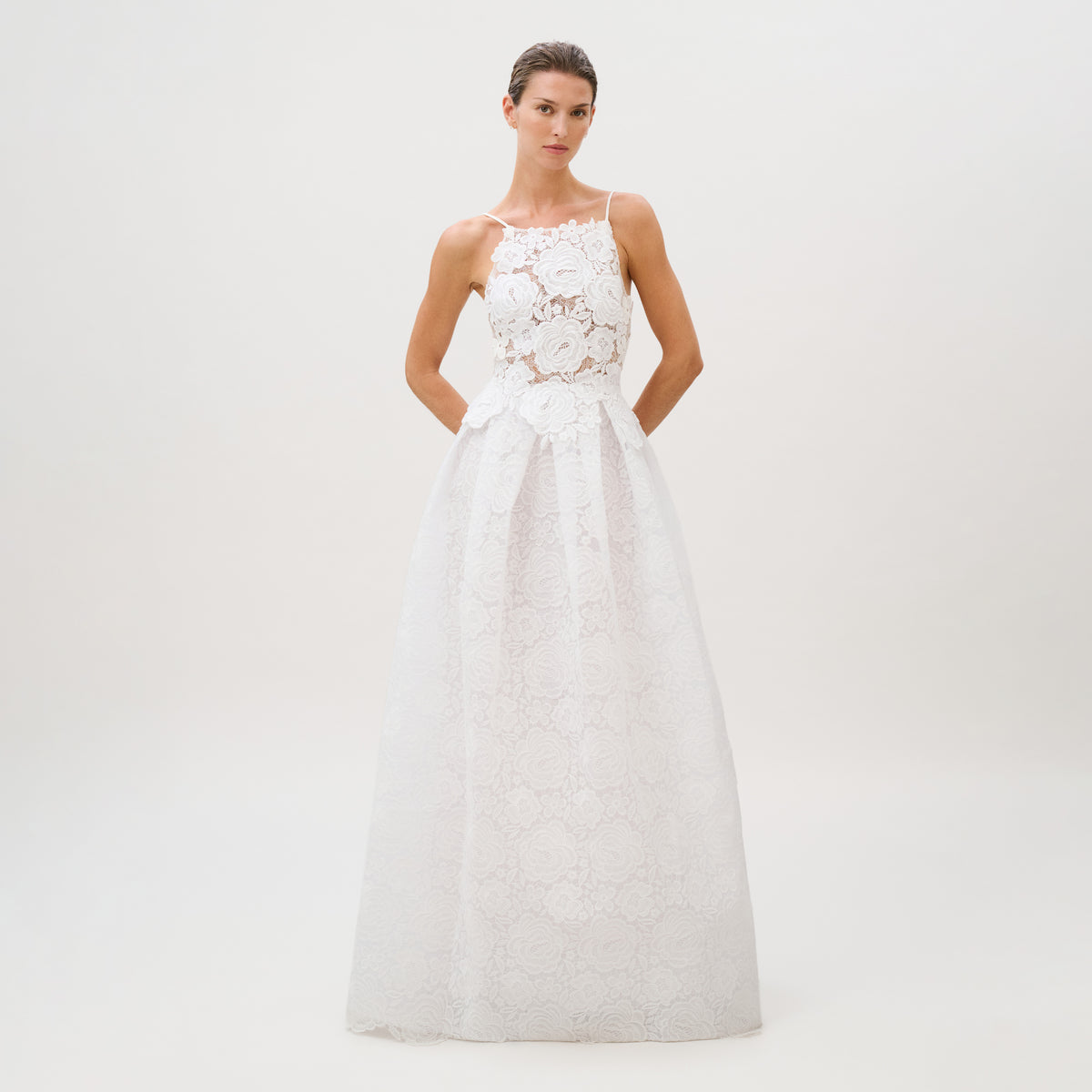 A woman models the Esme Dress, a sleeveless white lace bridal gown featuring floral patterns, a fitted bodice, and a full skirt. Made from luxurious Italian fabric in Colombia, she stands against a plain white background with her hands behind her back.