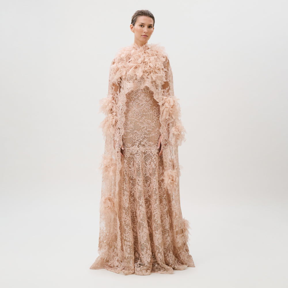 A person stands against a plain white background wearing the Eudora Dress, a floor-length gown in blush lace crafted from made in Italy fabric, paired with a coordinating feathered cape.
