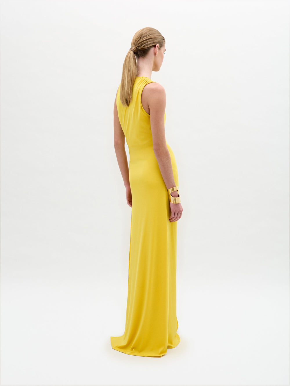A person models the Fadila Dress, a sleeveless yellow maxi with a fitted silhouette, deep V-neckline, and striking gold hip embellishment, standing against a plain white background.