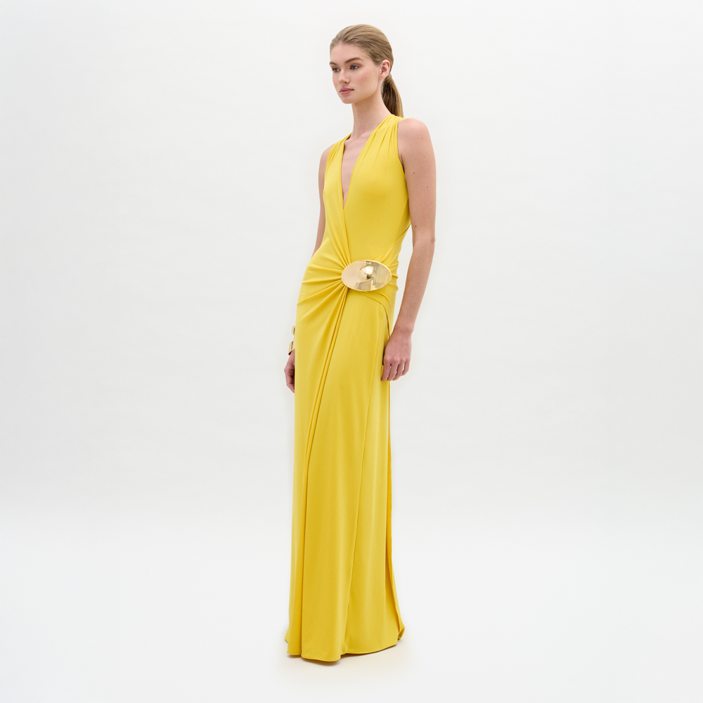 A person models the Fadila Dress, a sleeveless yellow maxi with a fitted silhouette, deep V-neckline, and striking gold hip embellishment, standing against a plain white background.