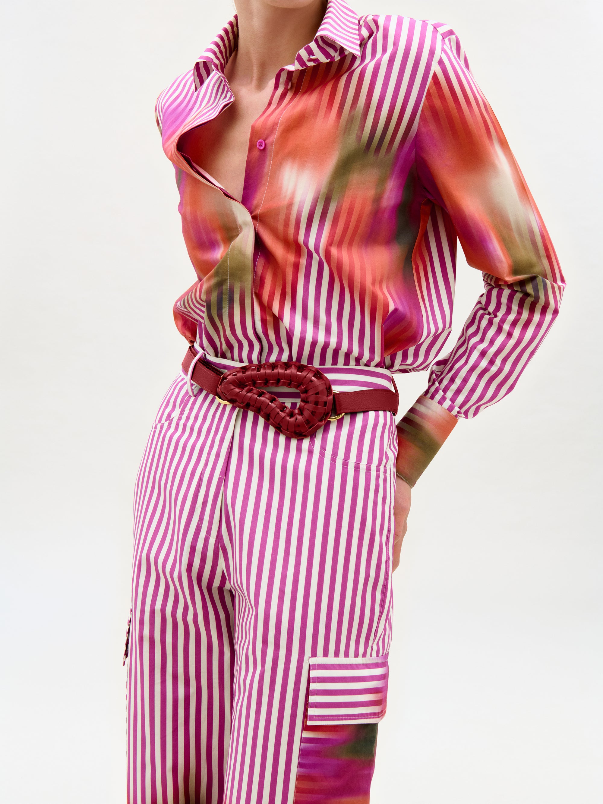 A person stands against a plain background wearing the Falak Pant—high-rise, straight leg pants with pink and white stripes—styled with a long-sleeve pink-orange gradient shirt and a braided red belt.