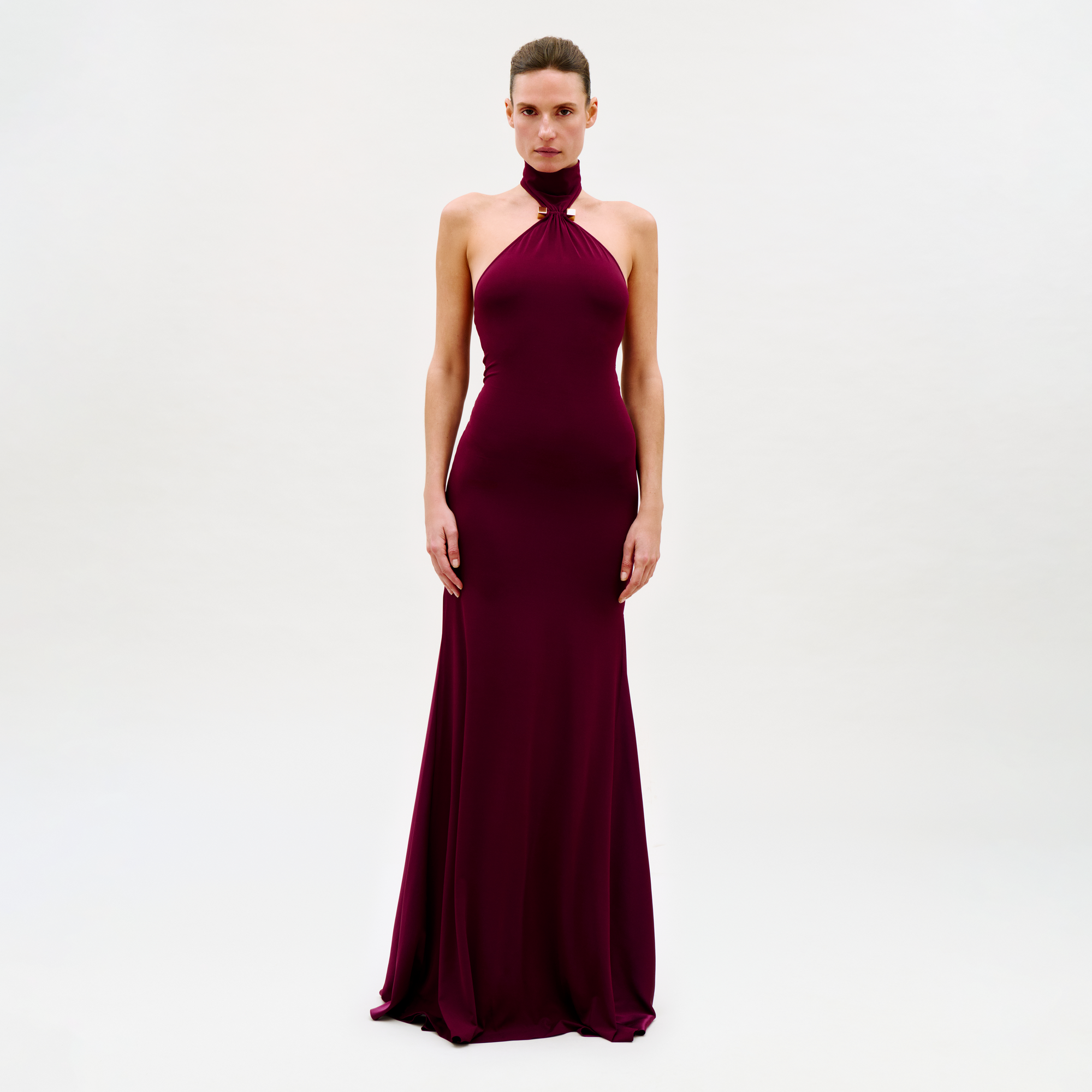A woman faces forward against a plain white background, wearing the Feist Dress—a long, sleeveless burgundy gown with a halter neckline.