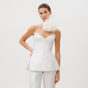 A woman wears the Filomena Top—a white, strapless, textured piece crafted from luxurious Italian fabric and finished with scalloped edges—paired with matching pants. She poses with a large white floral accessory on her shoulder against a plain light background, showcasing this ready-to-wear ensemble that exudes sophistication and timeless elegance.