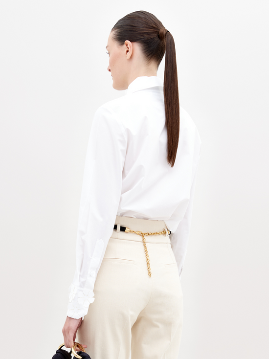 A person with dark hair tied back, wearing a Flower Blouse White with flower details on the collar and sleeves, beige pants, and a black belt with gold accents, holding a small black handbag. Pre-order now to ensure your outfit ships by November 15th, 2024.