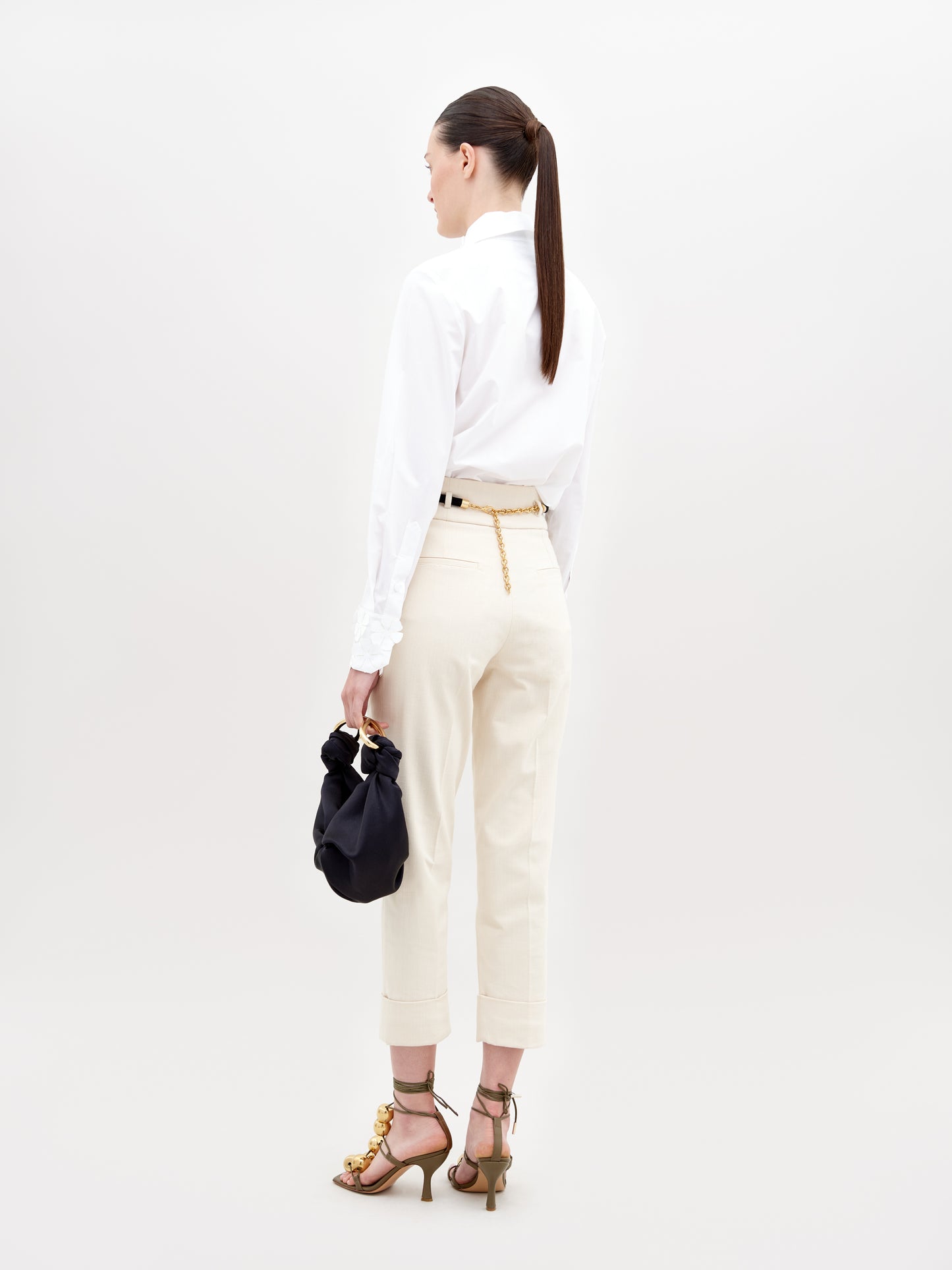 A person stands against a plain background, wearing a white blouse, Beryl Pant Beige, black belt, and golden embellished heels, holding a black handbag with a knot detail. This stylish ensemble is perfect for those who pre-order now to ensure it arrives by the November 15th, 2024 ship-by date.