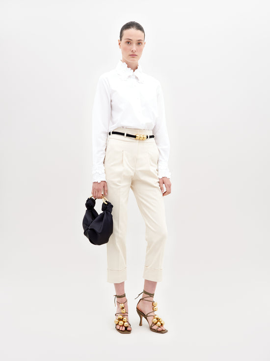 A person stands against a plain background, wearing a white blouse, Beryl Pant Beige, black belt, and golden embellished heels, holding a black handbag with a knot detail. This stylish ensemble is perfect for those who pre-order now to ensure it arrives by the November 15th, 2024 ship-by date.