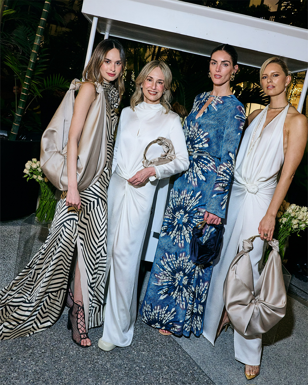 Four women in elegant dresses pose together at an event. Two wear white dresses, one a geometric pattern, and one a blue patterned dress. They hold large handbags and stand on a tiled surface.