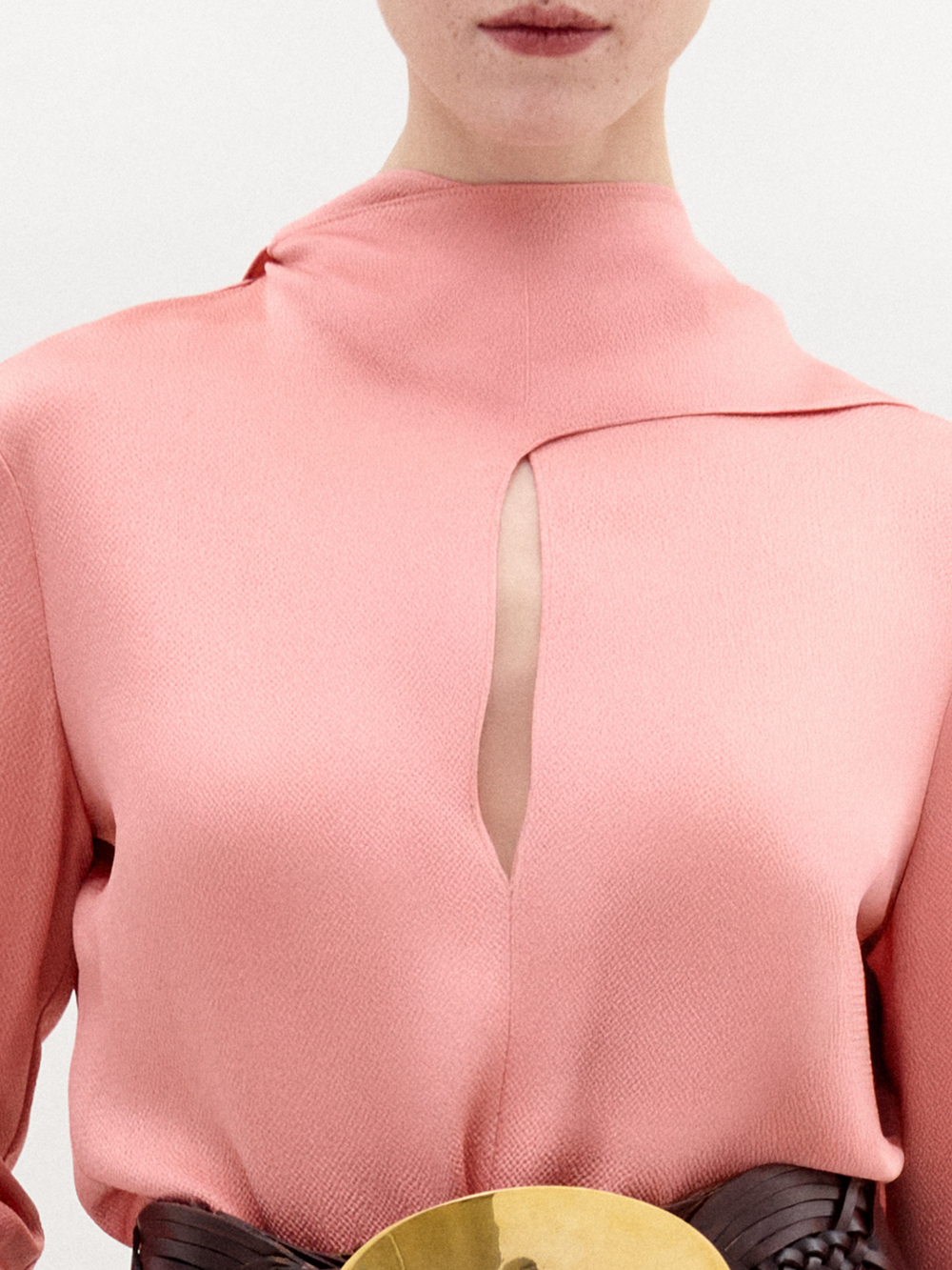 A woman stands against a plain background wearing the Gaby Blouse, a pink high-neck top with a front slit, paired with brown textured pants and a wide belt featuring a large gold oval buckle.