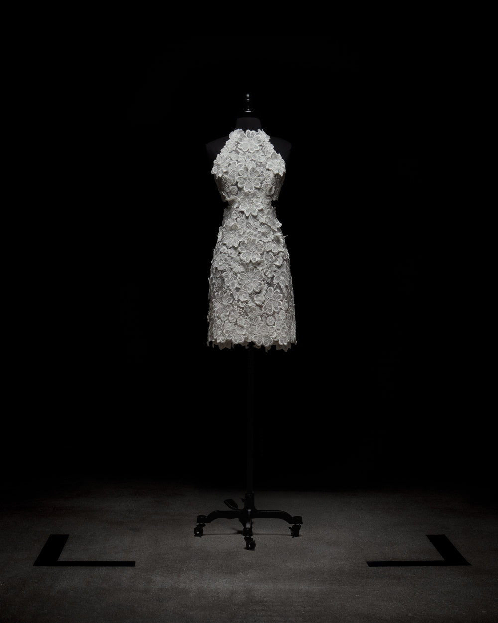 The Garazi Dress, a white sleeveless knee-length sheer lace dress with floral patterns, is displayed on a black dress form with a wheeled base against a dark background and sits on a gray floor.