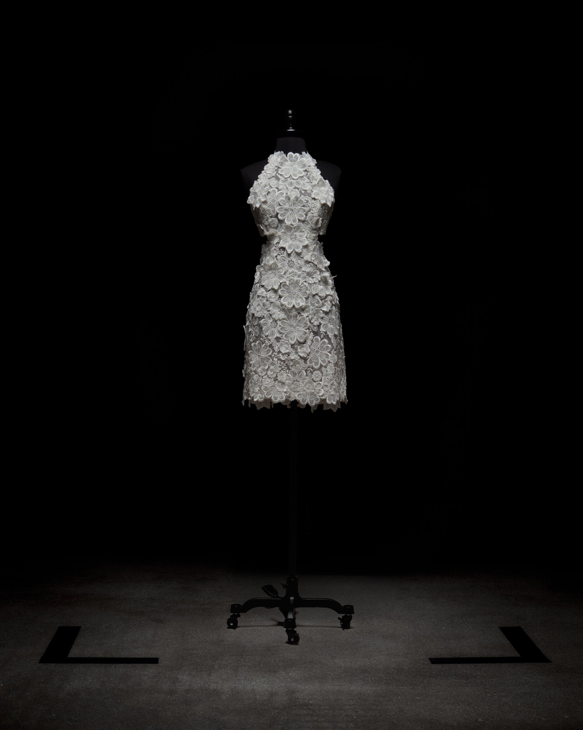 The Garazi Dress, a white sleeveless knee-length sheer lace dress with floral patterns, is displayed on a black dress form with a wheeled base against a dark background and sits on a gray floor.