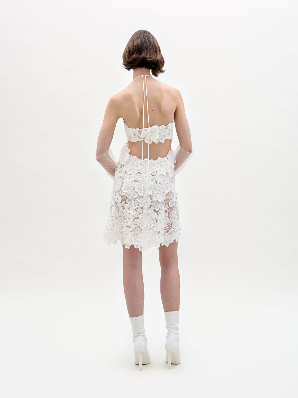 A person with short brown hair stands facing away, wearing the Garazi Dress—a short, white, sheer lace dress with floral patterns and an open back—styled with white socks and heeled shoes against a plain white background.