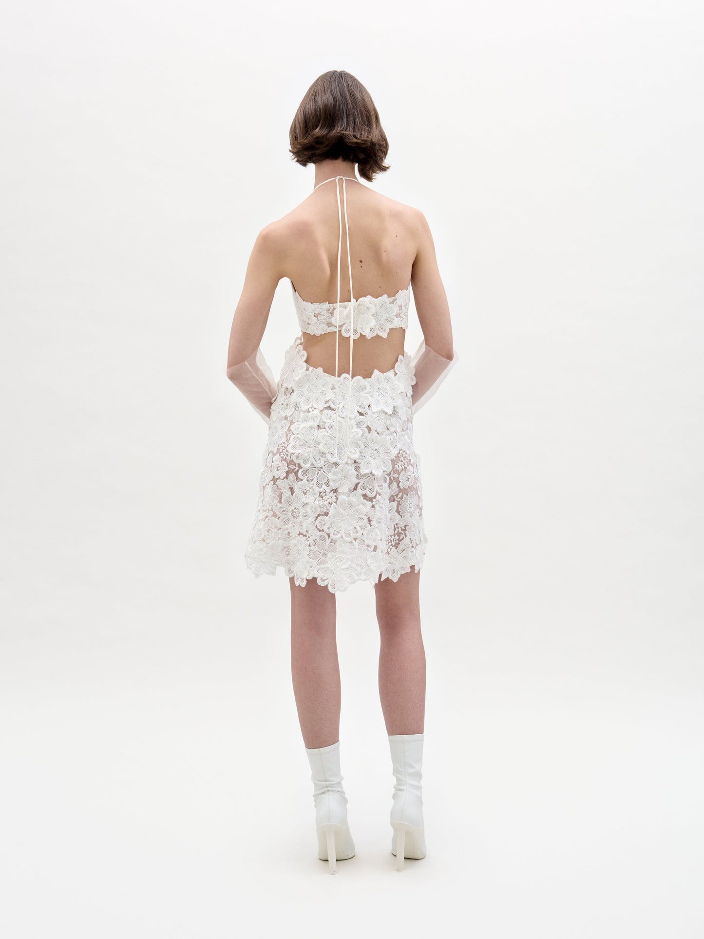 A person with short brown hair stands facing away, wearing the Garazi Dress—a short, white, sheer lace dress with floral patterns and an open back—styled with white socks and heeled shoes against a plain white background.