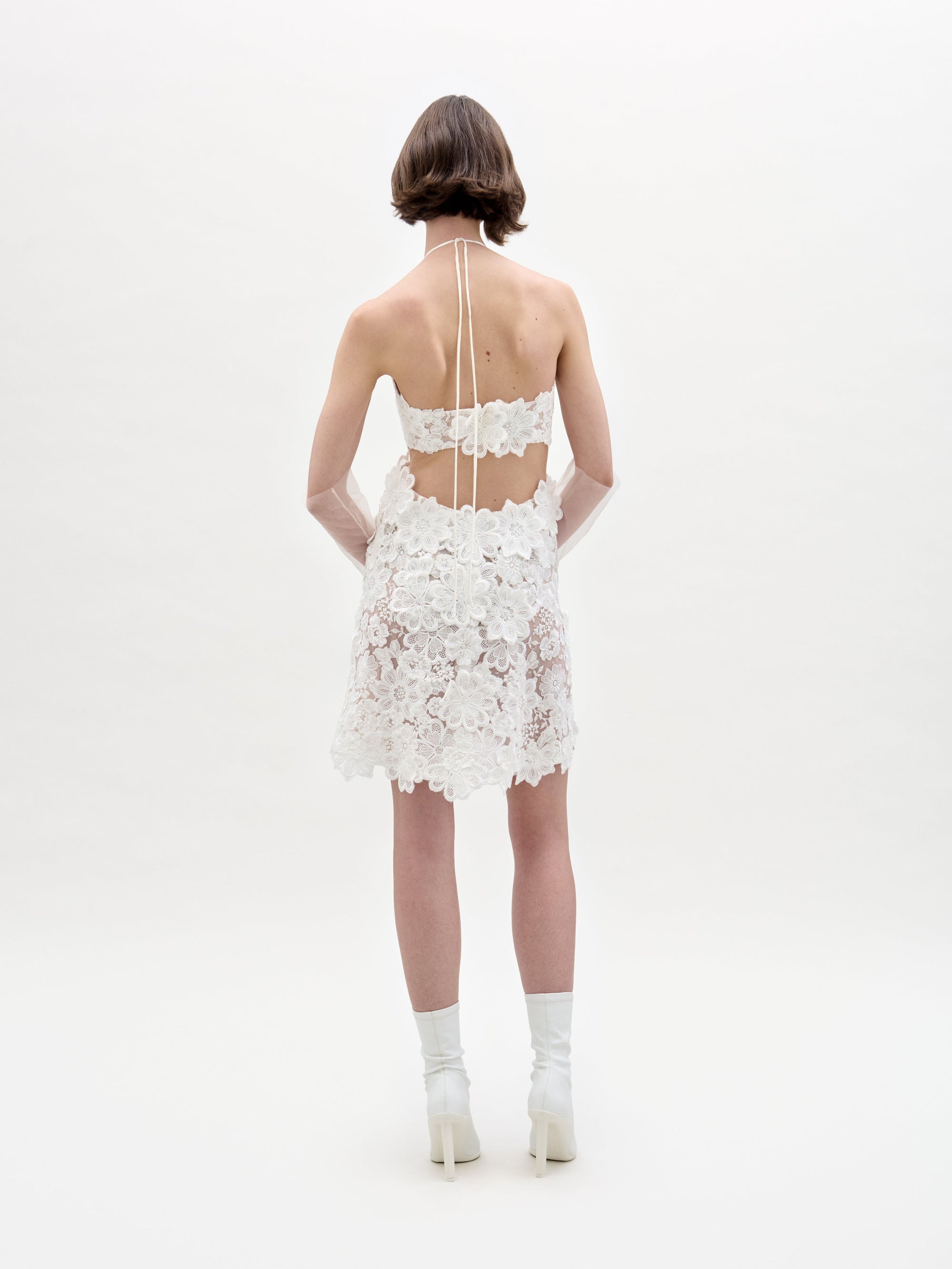 A person with short brown hair stands facing away, wearing the Garazi Dress—a short, white, sheer lace dress with floral patterns and an open back—styled with white socks and heeled shoes against a plain white background.