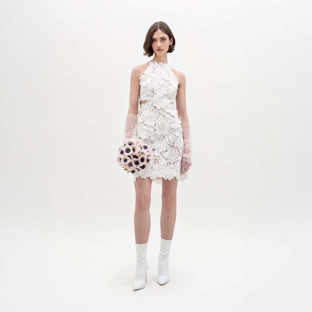 A woman wearing the Garazi Dress—a short, sleeveless white sheer lace dress—with sheer long gloves and white heeled boots holds a bouquet of light-colored flowers against a plain, light background.