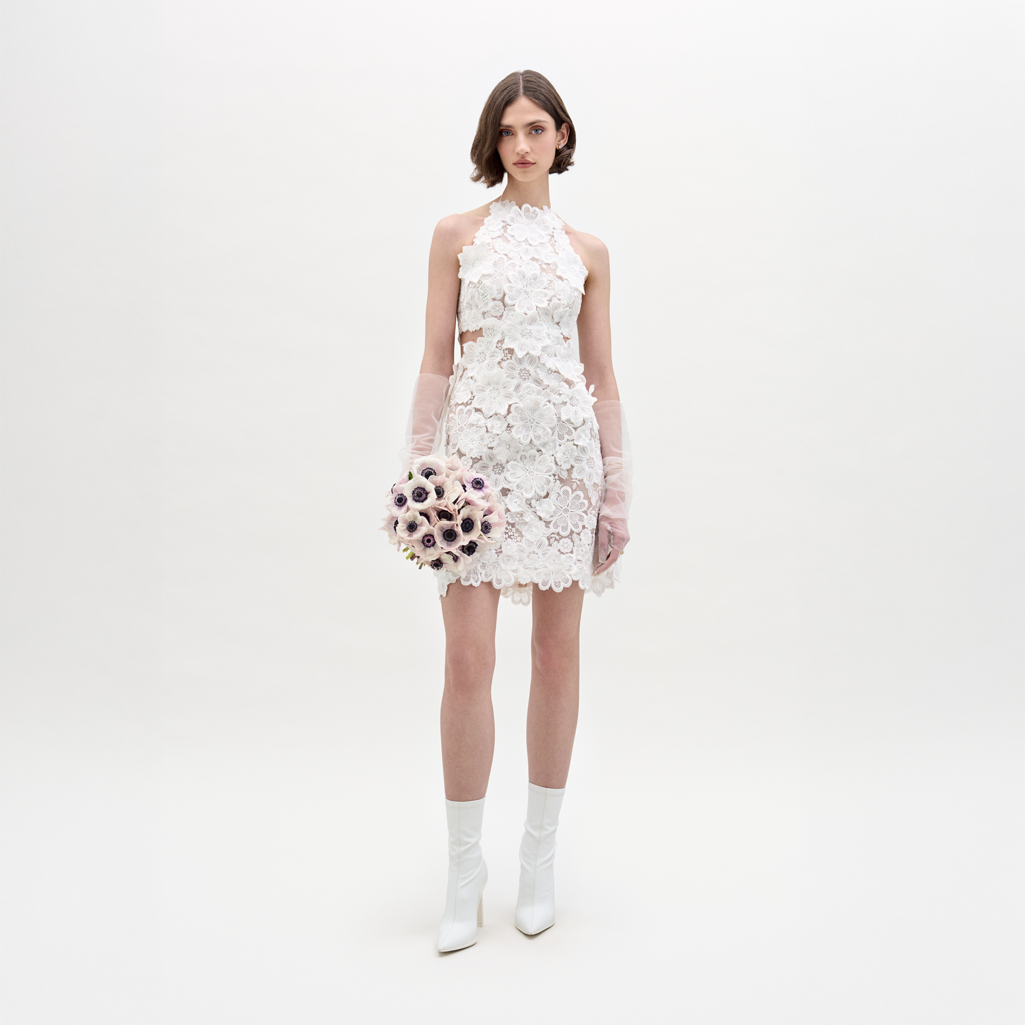 A woman wearing the Garazi Dress—a short, sleeveless white sheer lace dress—with sheer long gloves and white heeled boots holds a bouquet of light-colored flowers against a plain, light background.