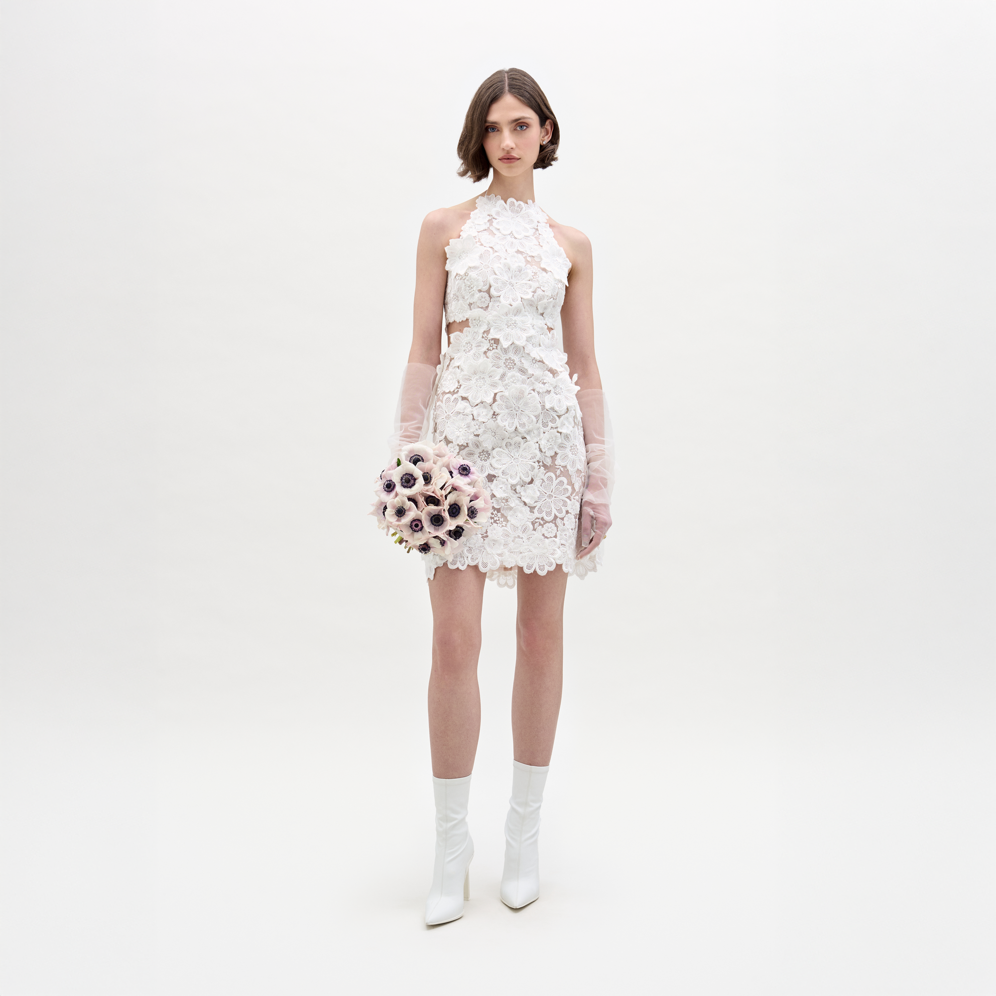 A woman wearing the Garazi Dress—a short, sleeveless white sheer lace dress—with sheer long gloves and white heeled boots holds a bouquet of light-colored flowers against a plain, light background.