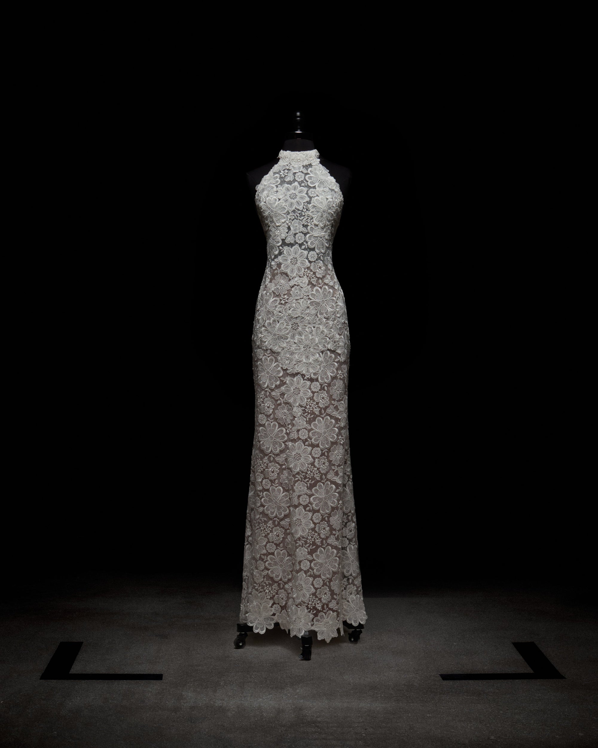 The Gardenia Dress is a floor-length, sleeveless sheer lace maxi dress with a high neckline and an intricate floral pattern, displayed on a black dress form against a dark background.