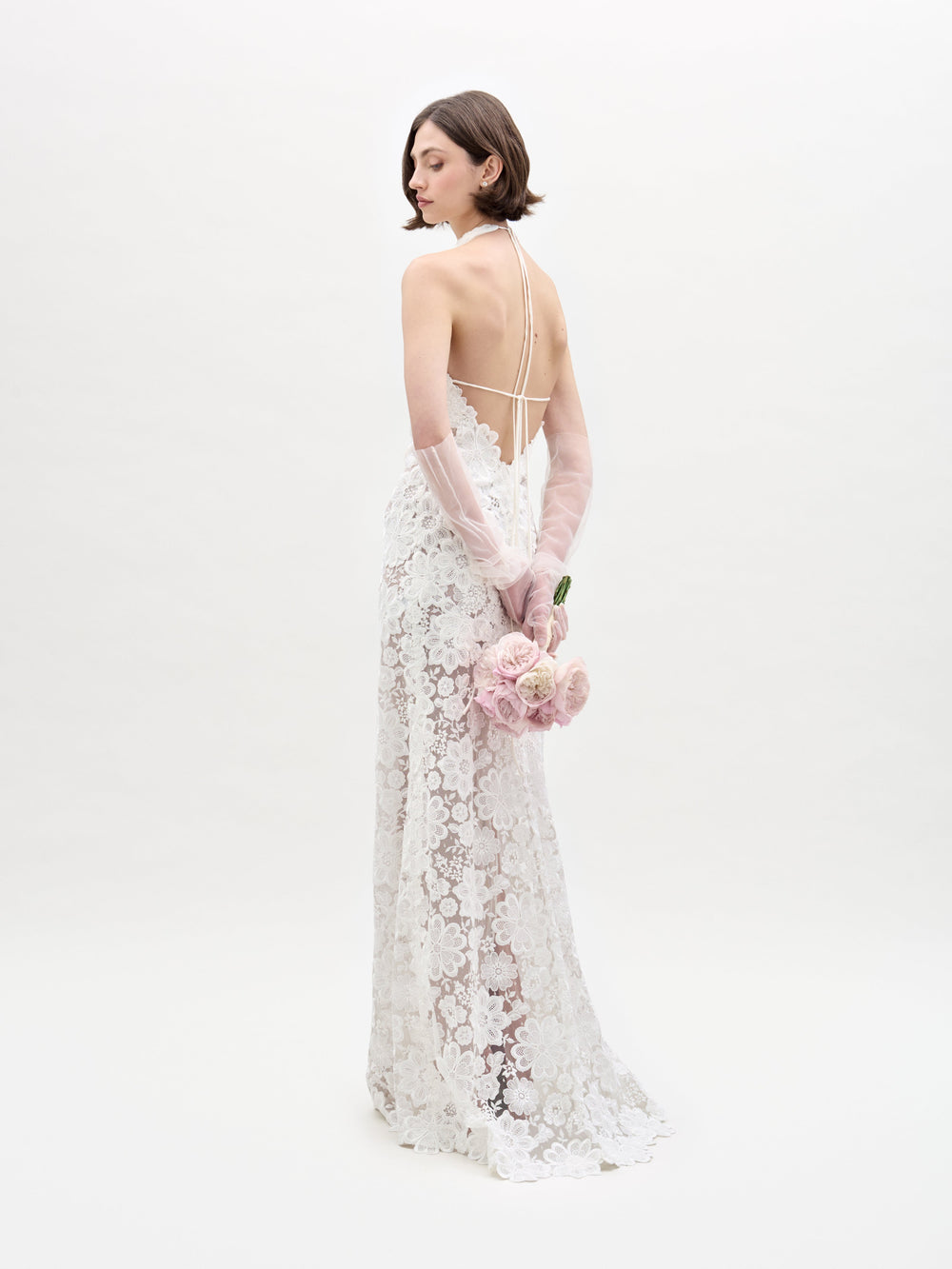 A woman in the Gardenia Dress, featuring sheer lace and a backless design, stands with her back turned and holds a bouquet of pale pink roses behind her against a plain white background.