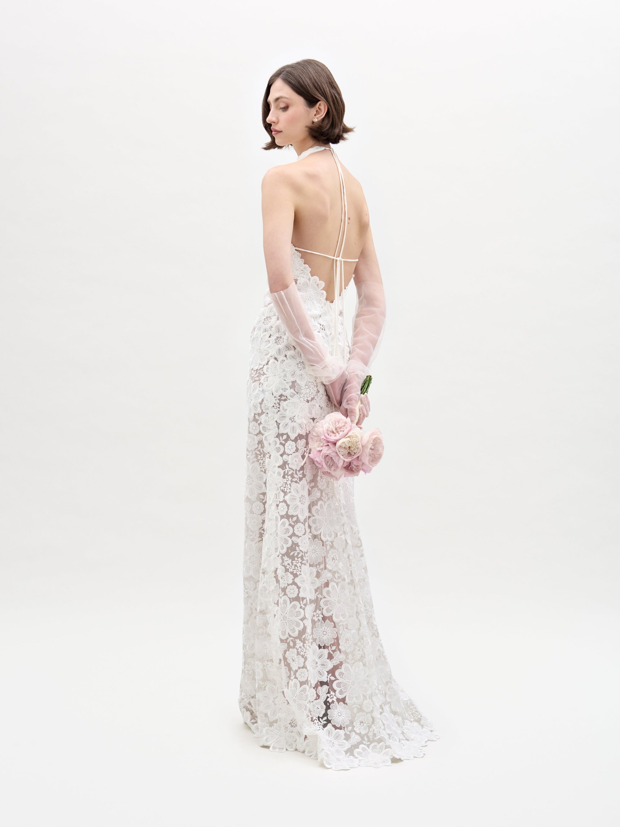 A woman in the Gardenia Dress, featuring sheer lace and a backless design, stands with her back turned and holds a bouquet of pale pink roses behind her against a plain white background.
