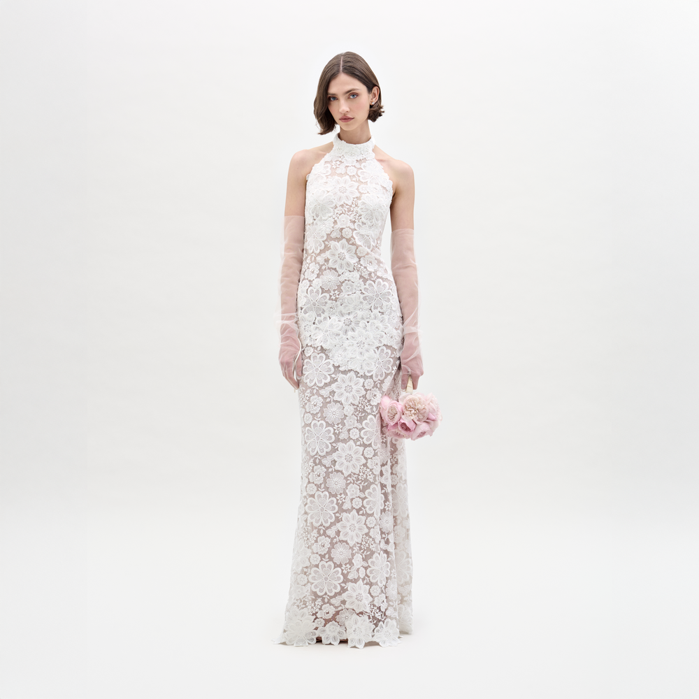 Wearing the Gardenia Dress, a sleeveless, floor-length sheer lace maxi, she holds pink flowers and dons sheer pink gloves, standing against a plain light background.