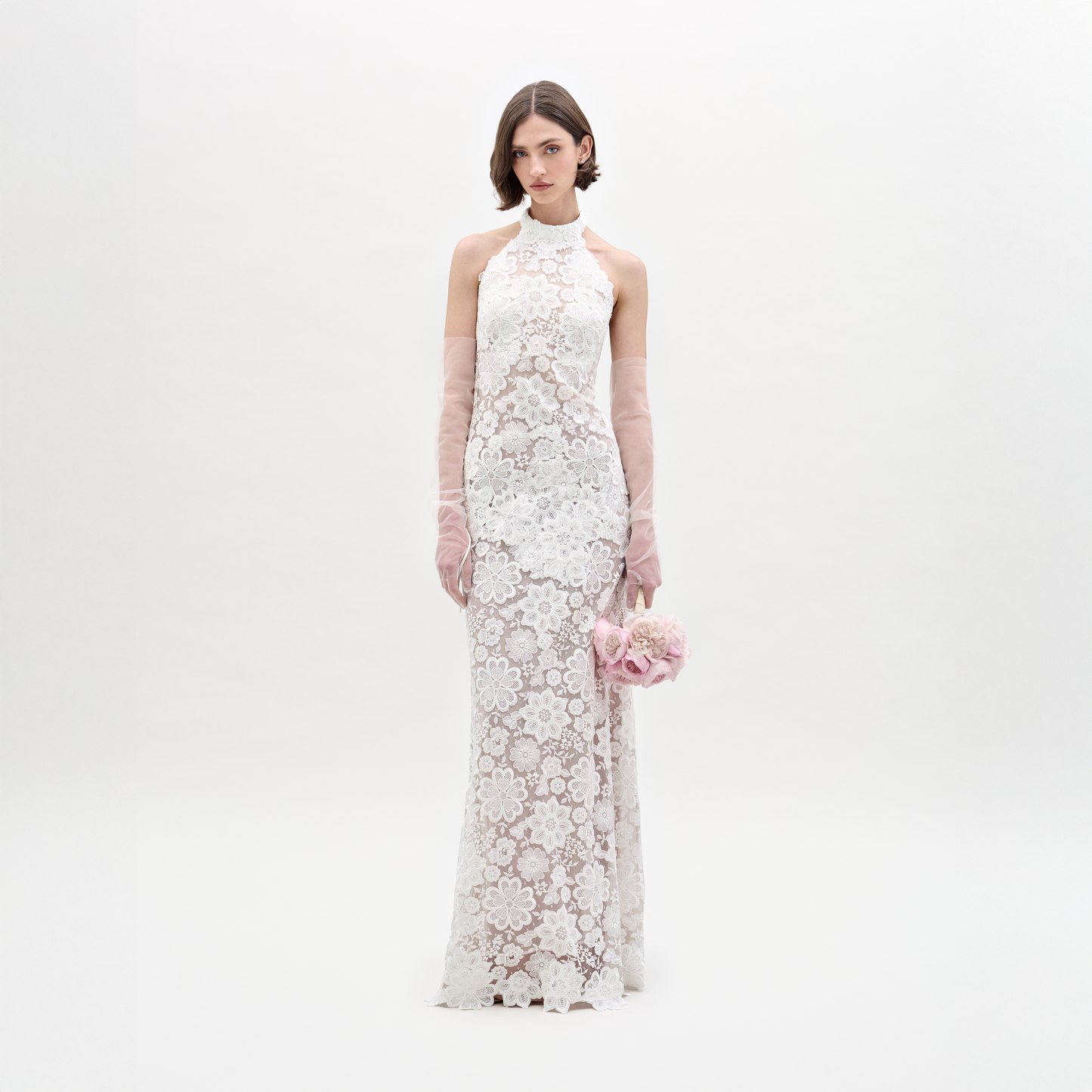 Wearing the Gardenia Dress, a sleeveless, floor-length sheer lace maxi, she holds pink flowers and dons sheer pink gloves, standing against a plain light background.