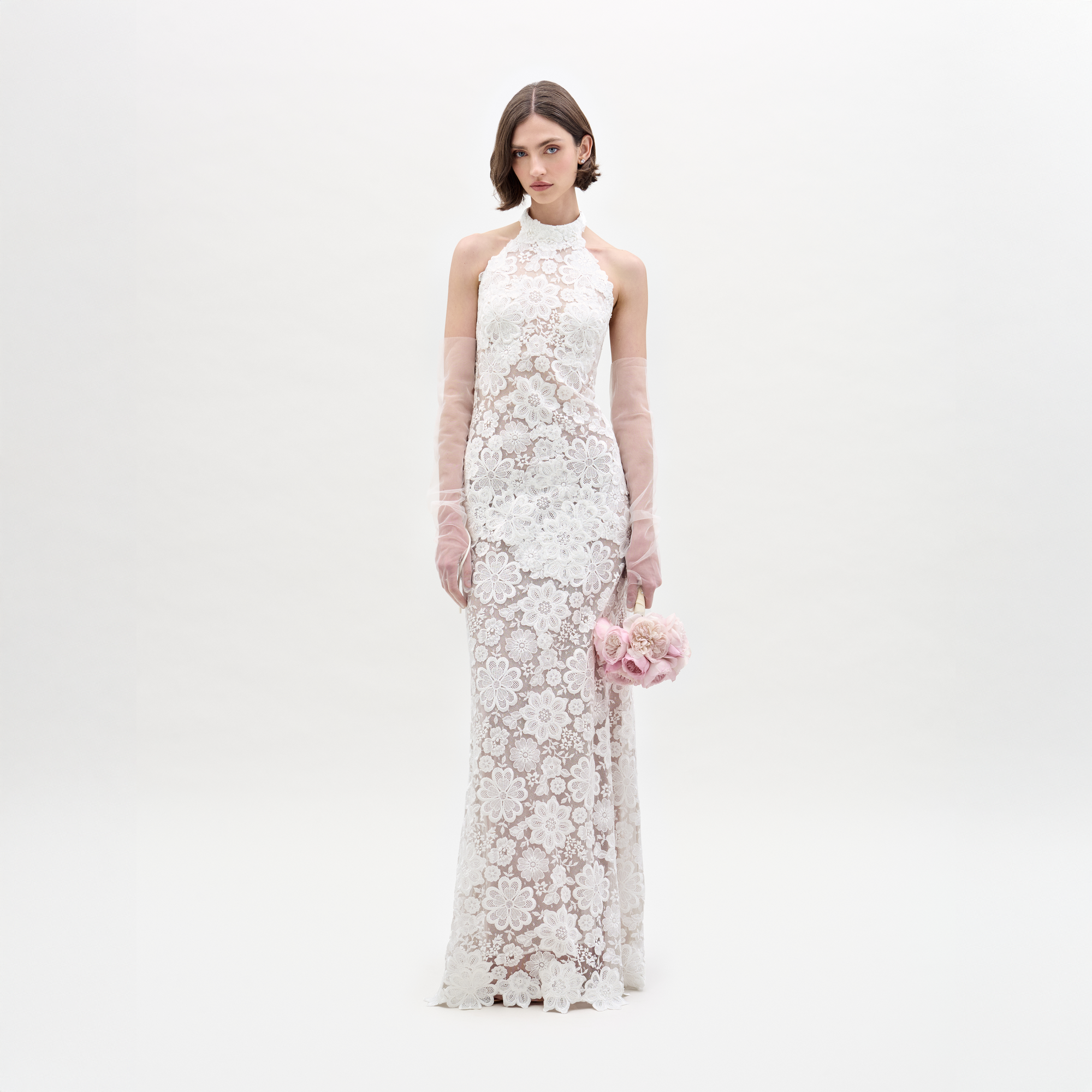Wearing the Gardenia Dress, a sleeveless, floor-length sheer lace maxi, she holds pink flowers and dons sheer pink gloves, standing against a plain light background.
