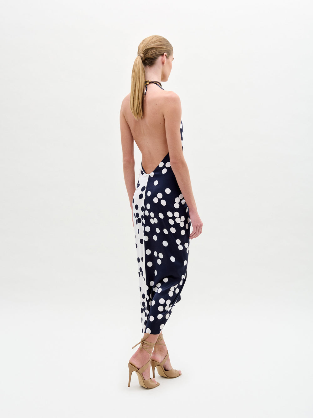 Wearing the Guadalupe Dress—a navy polka dot halter with a knotted front—paired with a colorful necklace and strappy heels, the person stands against a plain background.