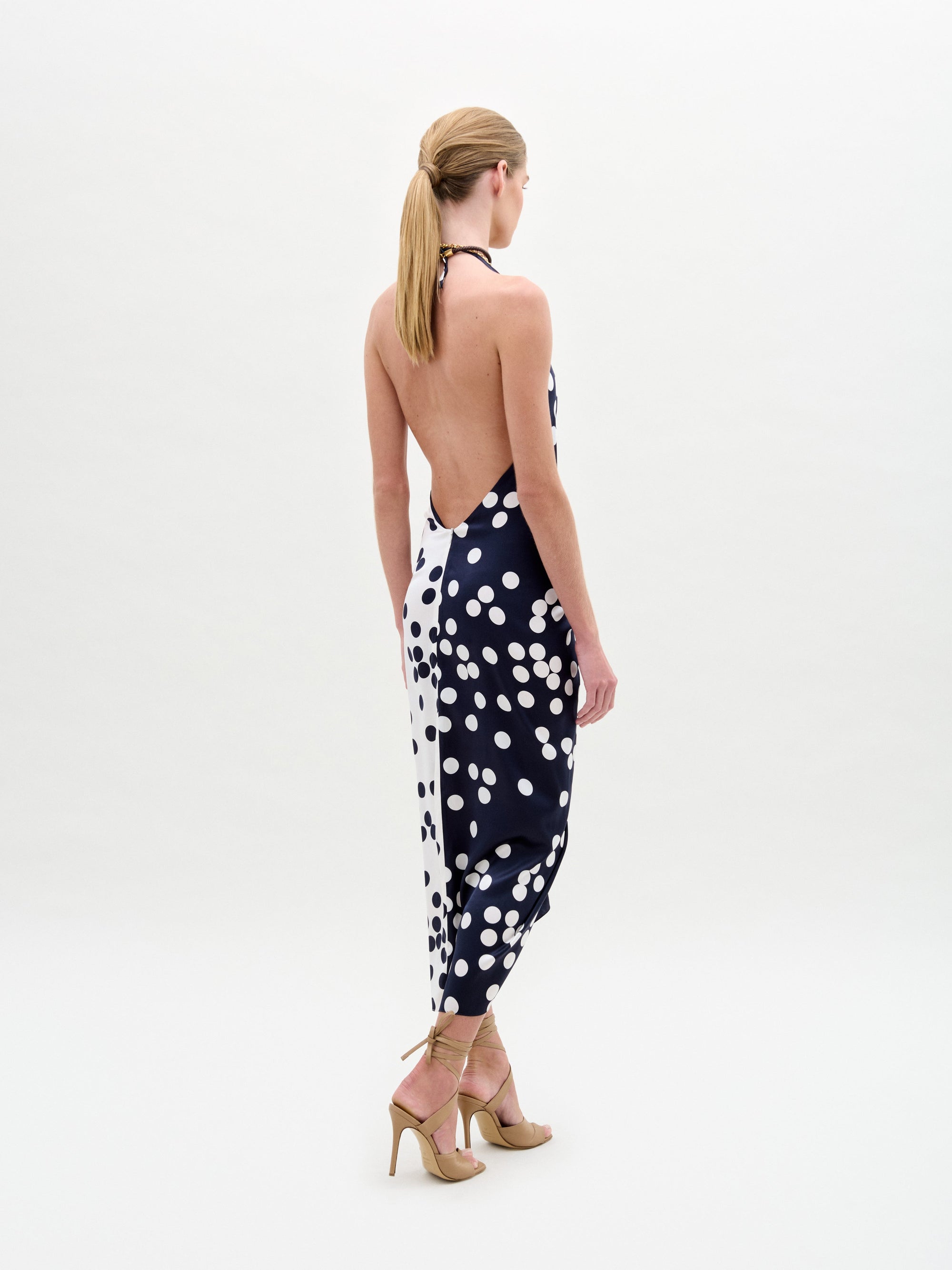 Wearing the Guadalupe Dress—a navy polka dot halter with a knotted front—paired with a colorful necklace and strappy heels, the person stands against a plain background.