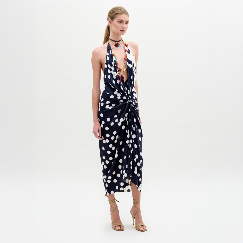 Wearing the Guadalupe Dress—a navy polka dot halter with a knotted front—paired with a colorful necklace and strappy heels, the person stands against a plain background.