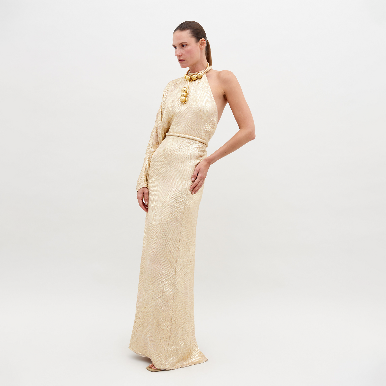 A woman stands against a plain background, wearing a Gael Dress Gold with a matching necklace and one sleeve. This exquisite ensemble is available for pre-order starting November 15th, 2024.