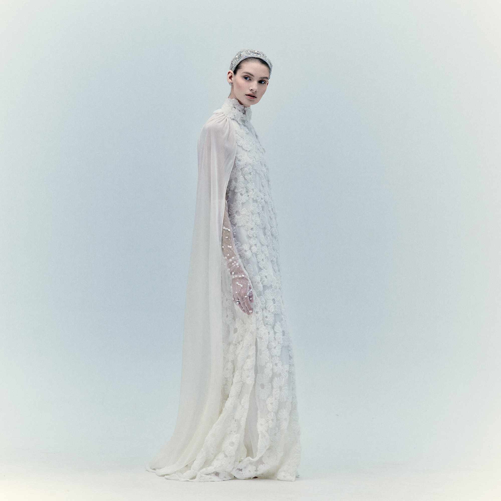 A woman stands against a pale background wearing the Gertrude Dress, a long, elegant white lace maxi with a high neck, sheer sleeves, cape, and matching headpiece and gloves adorned with delicate floral appliqué. She looks calmly at the camera.