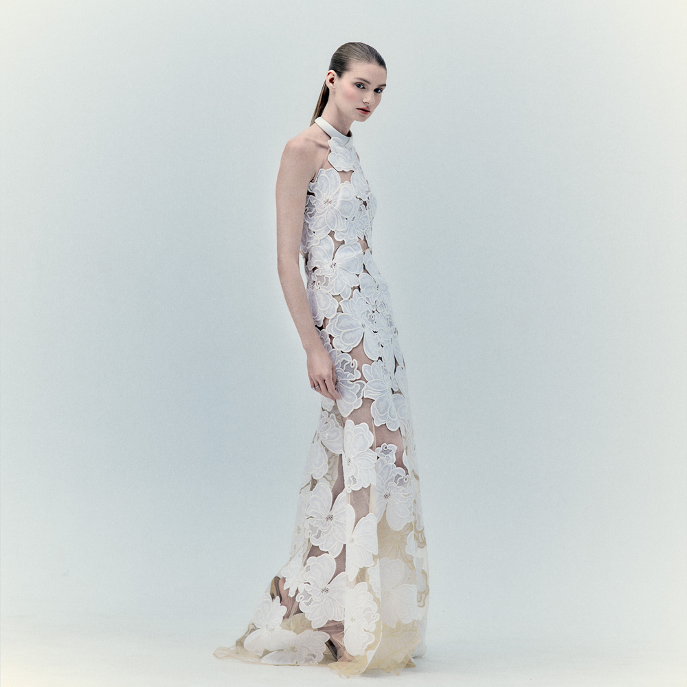Against a pale backdrop, a woman wears the Grace Dress—a long, sheer maxi adorned with large white floral appliqué. With slicked-back hair and a soft gaze to the side, she embodies elegance and an ethereal allure.