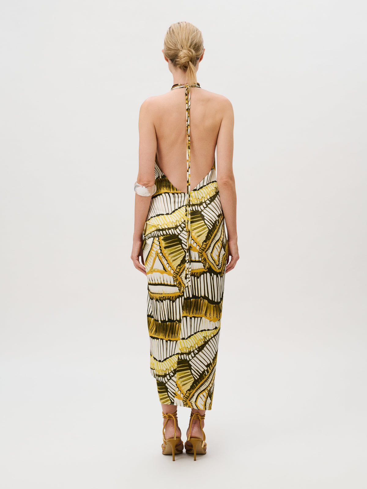 Back view of a person wearing the Guadalupe Dress, a sleeveless, backless midi dress of Italian fabric with a yellow and green geometric pattern, paired with strappy high-heeled sandals.