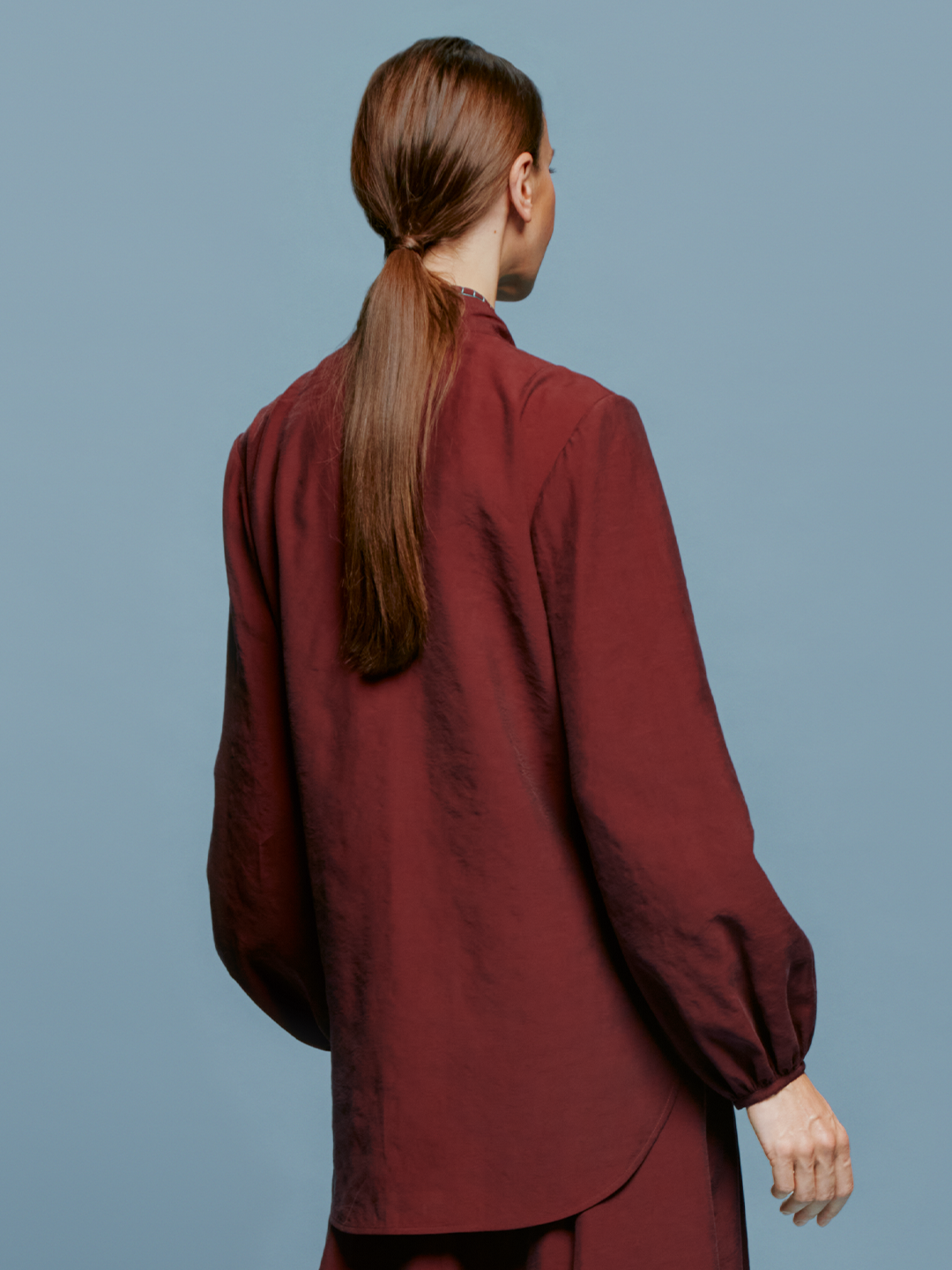 A woman with brown hair is wearing the Honey Blouse Burgundy, a wrap-style top with balloon sleeves, standing against a blue background, ready for the September 2024 launch.