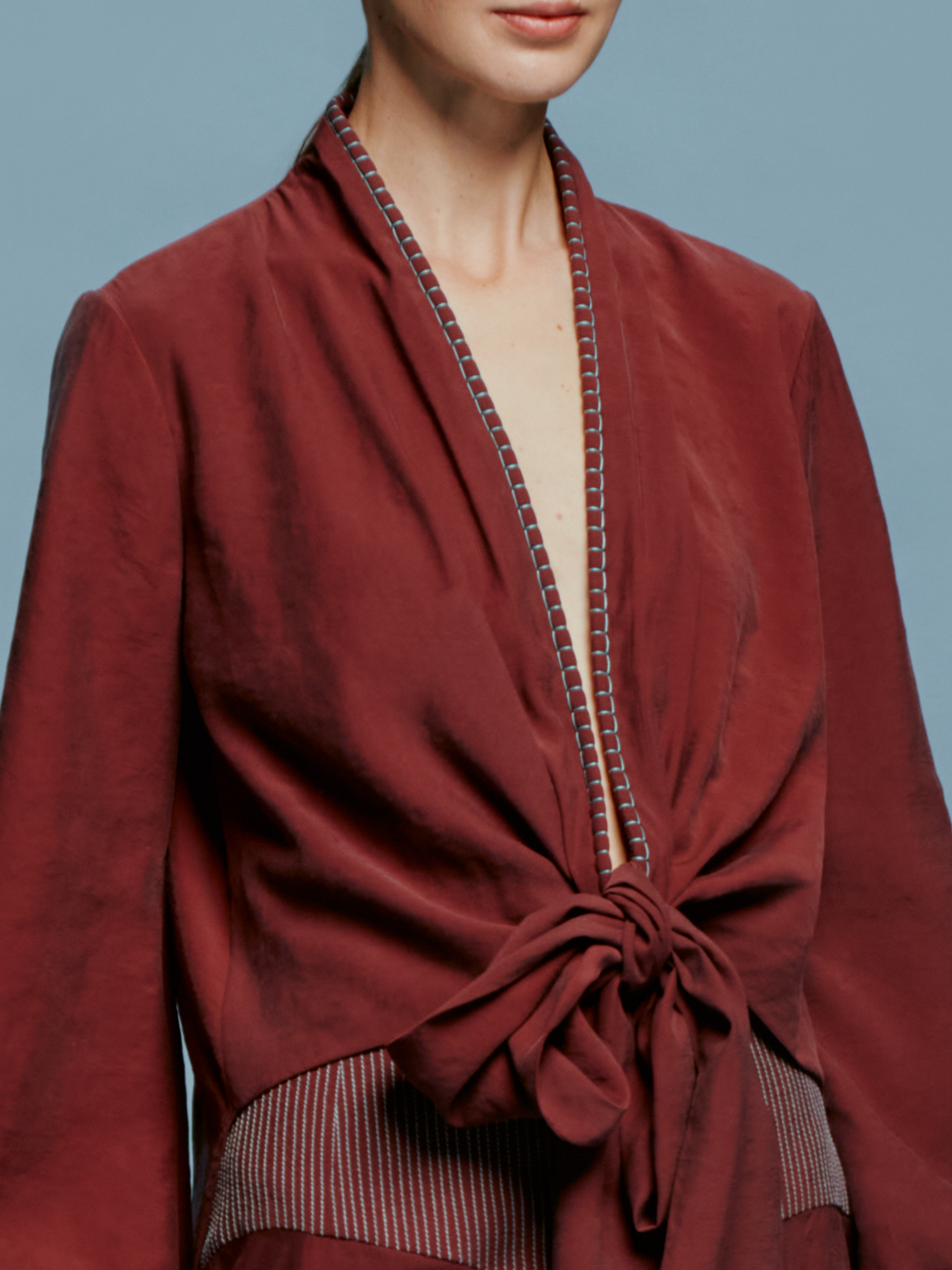 A woman with brown hair is wearing the Honey Blouse Burgundy, a wrap-style top with balloon sleeves, standing against a blue background, ready for the September 2024 launch.