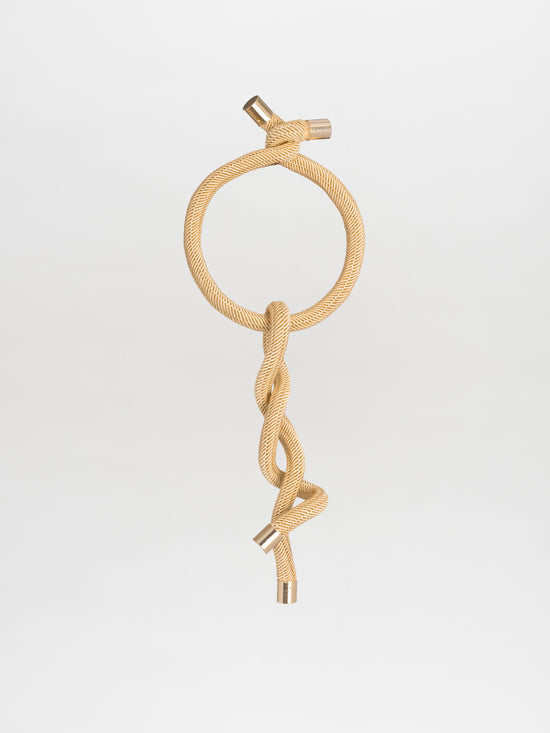 The Hazel Necklace Gold features a circular loop with an interwoven design, embellished with viscose threads and metallic accents at the end caps, all set against a plain background.