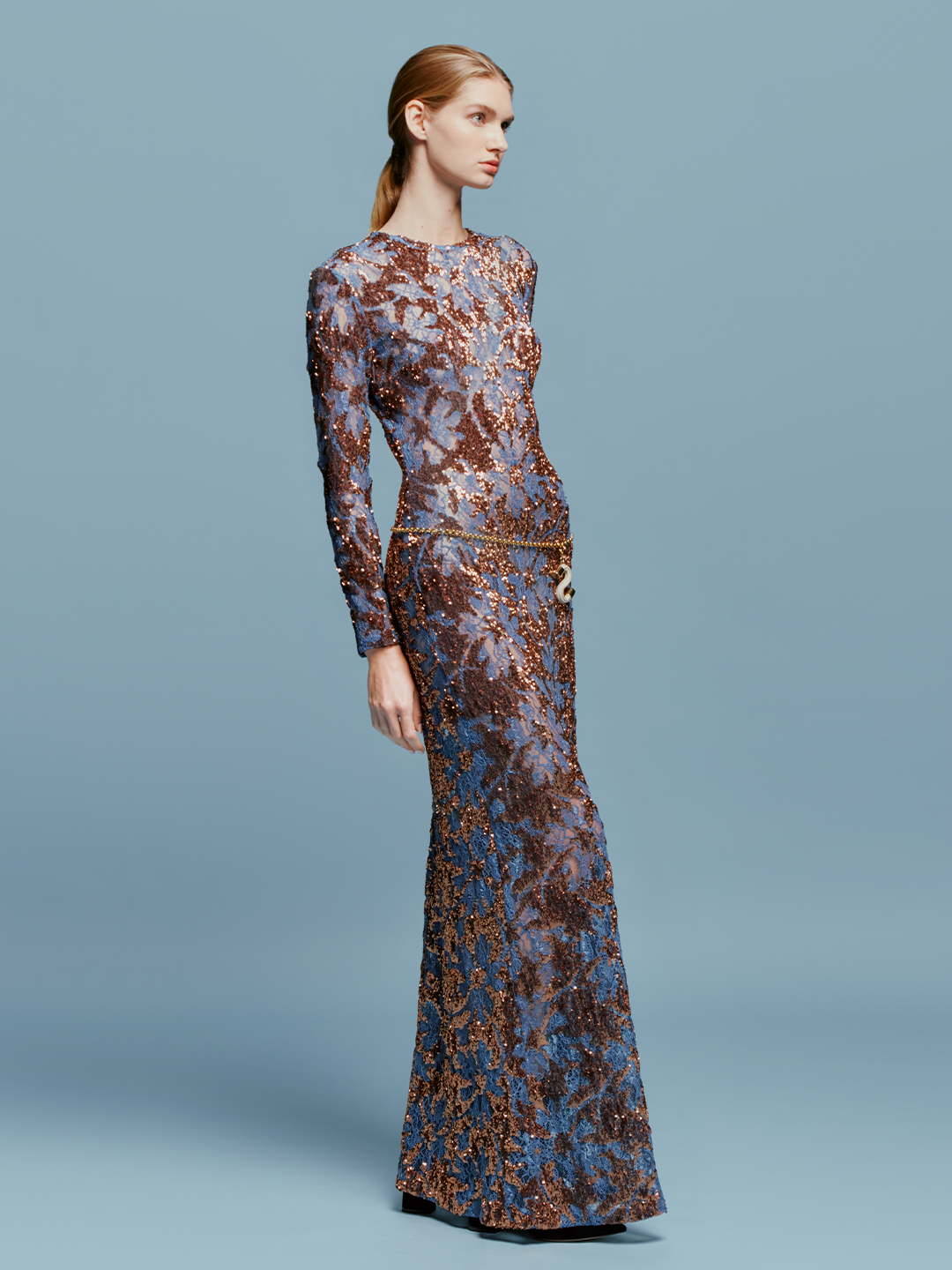 Person in the Ida Dress Navy Ochre Sequin, a long-sleeved, floor-length gown featuring an intricate blue and brown pattern, stands against a plain blue background, ready to dazzle at the gala on September 2nd.