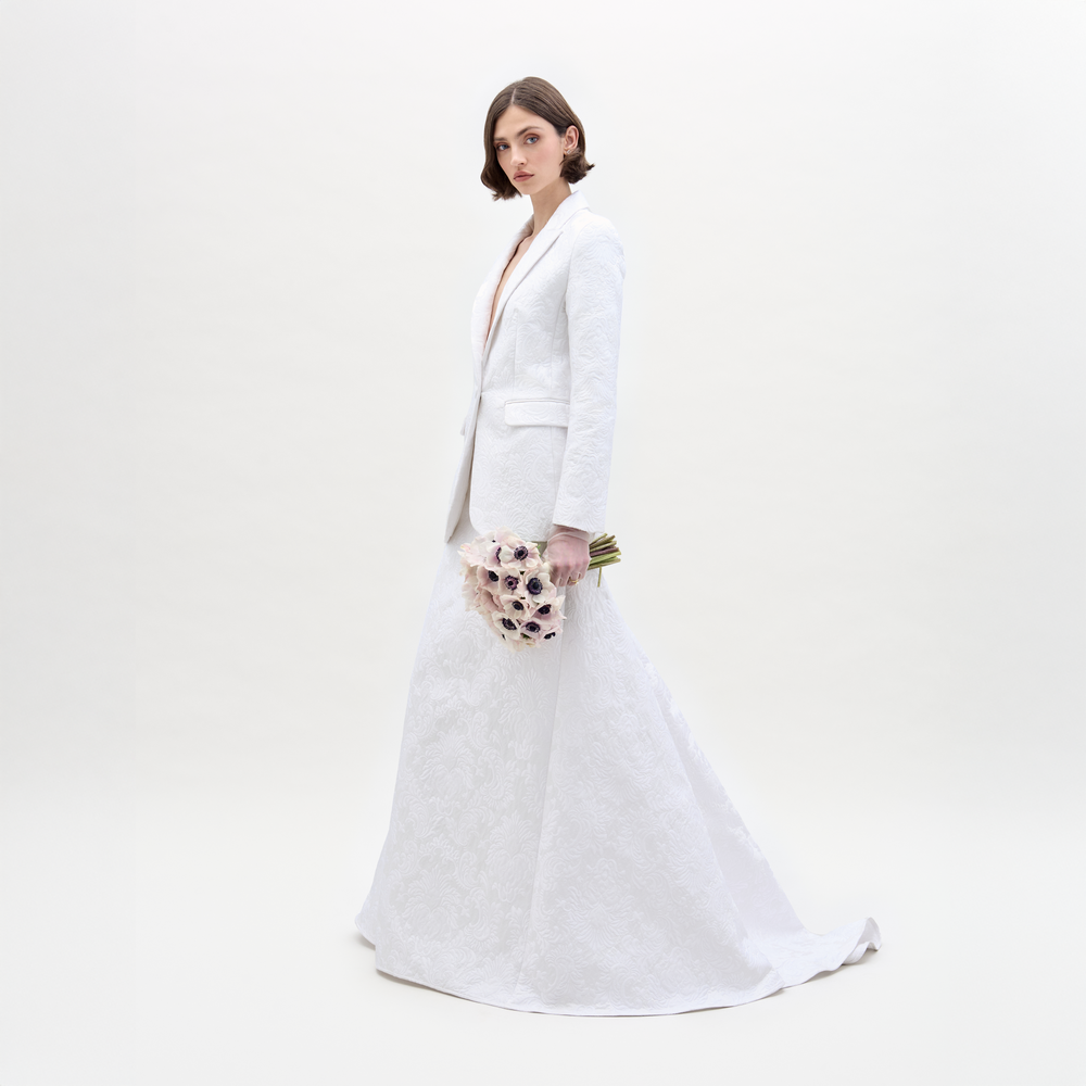 A woman with short brown hair wears the Imola Skirt and a matching white blazer, holding a bouquet of light flowers. She stands against a plain, light background.