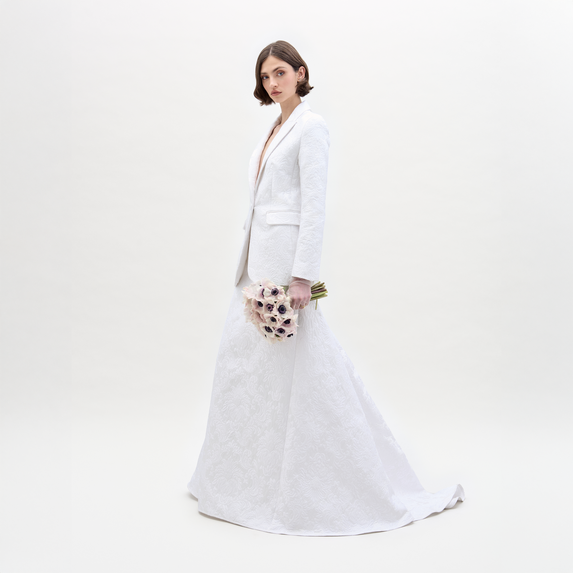 A woman with short brown hair wears the Imola Skirt and a matching white blazer, holding a bouquet of light flowers. She stands against a plain, light background.